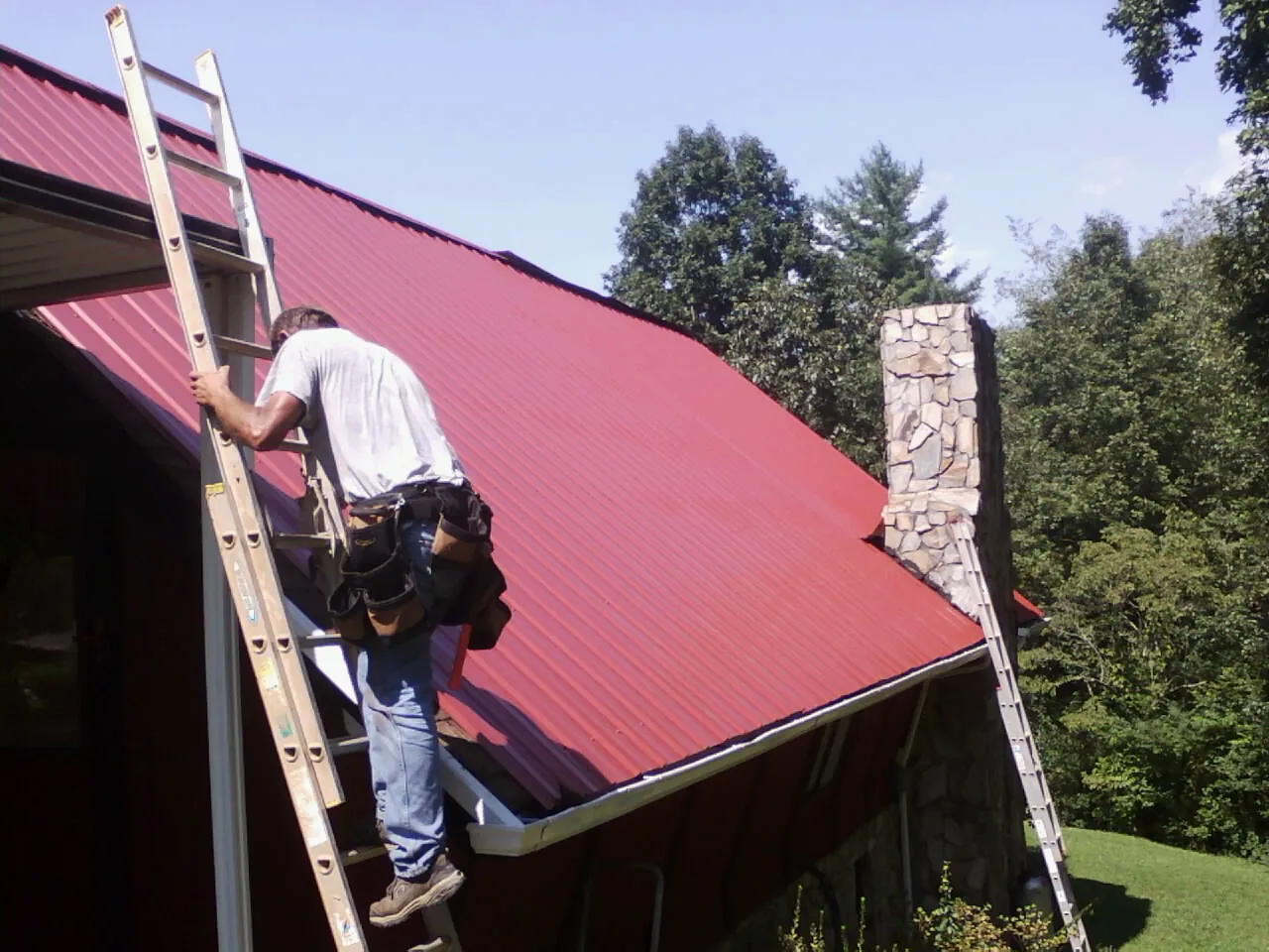 Roofer on a ladder beside a red metal roof and stone chimney — Soffit & Fascia Repair by Roofing Brothers in Peoria Heights